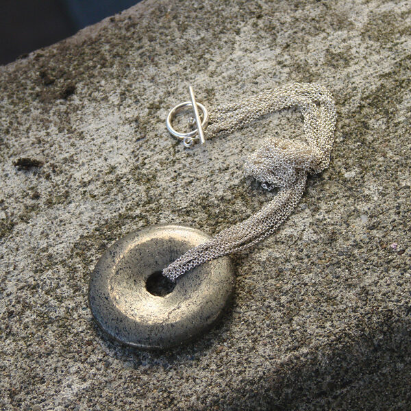 A unique silver piece, handcrafted by Minda Lane, delicately hangs from a chain on a sleek concrete wall.